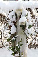 Classical female statue with ivy stole laden with snow against Ligustrum vulgare 'Atrovirens' in winter - Welsch Garden, Berlin, Germany