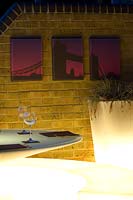Contemporary lit plastic dining furniture with brick wall and canvas prints. Container with ornamental grass. Wapping Balcony
