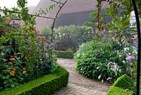 Metal arch with annual climbers leads into exotic style parterre with lox clipped box hedging, brick and tile path. Black Essex barn in background. 