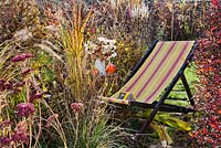 Deck chair amongs perennials and grasses in autumn. Pennisetum, Crocosmia 'Lucifer', Aster, Sedum, Panicum virgatum.