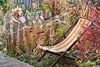 Deck chair amongst perennials and grasses in autumn: Pennisetum, Crocosmia 'Lucifer', Aster, Sedum, Panicum virgatum.