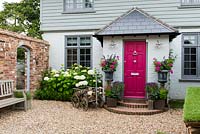 Cottage with front door flanked by pair of urns planted with Fuchsia 'Sunray', Salvia 'Black Knight', Pelargonium hortorum 'Orbit Synchro' and pink cosmos. Left bed planted with Hydrangea 'Annabelle' and box hedge. Doorway in brick wall on left leads into small courtyard garden.