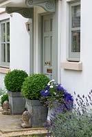 Metal containers with topiary box balls beside a front door. Lavender fills a gravel bed leading along the side of the house.