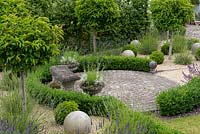 A circular patio with stone bench surrounded by low box hedging, portuguese laurel standards and stone spheres.