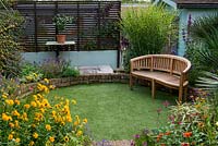 A sunken circular lawn of artificial grass, with a wooden bench in front of miscanthus sinensis 'Zebrinus'.