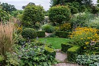 A suburban garden with a circular structure created by shaped box and a clover lawn. Two Photinia x fraseri standards divide the garden and provide height. Deep borders of mixed planting includes Astrantia, Rudbeckia, Salvia, Verbena and ornamental grasses.