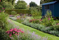 A grass path running between a pink and purple double border planted with Nepeta 'Six Hills Giant', Aster 'Jungfrau', Salvia bethellii, Dahlia 'Purple Haze', Sedum 'Autumn Joy', Buddleja and Cosmos 'Dazzler'. A summer house called the Shepherd's Hut