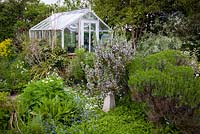Greenhouse with herb garden in front. The stone sundial is overshadowed by mature Rosmarinus officinalis - rosemary. Other herbs include Saponaria officinalis - soapwort, marjoram, Sanguisorba 'Tanna', hyssop, sweet cicely, white-flowered sage and phlomis.