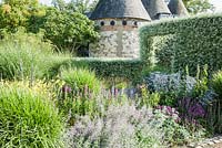 The Courtyard Garden designed by Piet Oudolf and John Coke features a circular entrance of clipped silver pear, Pyrus salicifolia 'Pendula', here seen against a backdrop of oasthouses, and behind a bed of grasses and perennials including eryngiums, stachys, hemerocallis and lythrum. Bury Court Barn, Bentley, Hants, UK