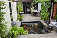 Outdoor dining area on a small patio with pergola covered with Parthenocissus. Agapanthus orientalis White in pot. Carpinus betulus - Hornbeam in modern pots. Pond with Koi Carp. Family Fabry - Mathijs. Belgium
