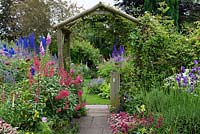 A cottage garden with path leading to rustic wooden arch, inbetween borders of phygelius, cosmos, penstemon, campanula, delphinium, foxglove, thalictrum, viola, valerian, lavender and astrantia.