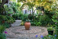 Circular courtyard made from circle of brick filled in with gravel, a terracotta urn at its centre. Surrounding borders planted with shade-loving hellebores, hardy geranium, dicentra, ferns, lamium and foxgloves. Behind on right, Japanese maple overhangs seat. 