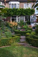 A suburban garden with a circular structure created by shaped box and paving. Deep borders of mixed planting include Geranium, Alchemilla, Astilbe, Salvia and ornamental grasses.