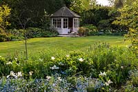 A sunny spring garden with mixed border of Forget-me-nots and white Tulipa Purissima. Behind the lawn sits a small wooden summerhouse and stone patio.