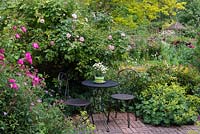 A secluded seating area on a brick patio surrounded by alchemilla, poppies and roses 'Vanity', 'Blush Noisette'.