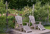Two ariondack chairs on a raised deck in front of 'Bessie's End' - an informal parterre with wildflowers, lupins and Rosa 'Mme Alfred Carriere' weeping standards.