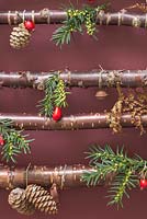 A hanging Christmas tree made with Birch branches, against a red background. Decorations feature Pine cones, feathers, Rose hips and Pine foliage