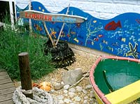 School playground garden - lobster lagoon.  Designer: Yvonne Mathews. Close up of small boat shingle and pebbles. Lavender  - lavendula planting, decking and ocean life mural in shape of wave and orange buoy.