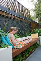 Girl sitting on timber built-in bench reads in inner city courtyard garden. Various shade loving plants seen. 