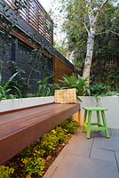 Timber bench and raised concrete planter in inner city courtyard garden. Planter contains Philodendron 'Xanadu' and Renga lilies. Camellias along wall. Under bench lighting illuminates maiden hair fern