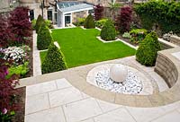 Contemporary urban garden on three levels featuring: marple sphere and pebble fountain and paved terrace, lawn, box pyramids, tulips and maples, basement seating area and conservatory