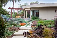 View across concrete path and mixed beds to modern house and outside eating area.  Debora Carl's garden, Encinitas, California, USA. August.