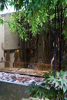 Black bamboo stems of Phyllostachys nigra and small acer growing in front of the patterned concrete-rendered low wall with water feature. Gold metal sheeting hung on back wall. 