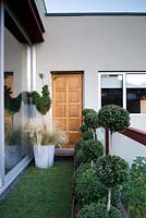 Balcony with glass door on the left and glass baluster on the right, astroturf flooring and box topiary in containers. One galvanised bin container with stipa grass. 
