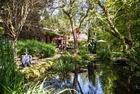 House, terrace with garden furniture, parasols and pond with waterside planting - July, Les Jardins de la Poterie Hillen