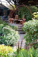 View over planting down garden with paving path, with steps, ornaments, rusted steel objects, ferns, phlomis, crambe maritima, seats and a summer house in the background. Rusted steel rod arch. 