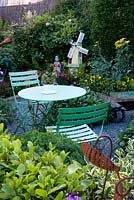 Garden packed with plants, ornaments and found objects including a windmill, boy figure, metal fish, wheelbarrow. Slate chip path. Cafe table. Colourful planting. 