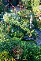 View from above of densely planted garden with plants for contrasting green foliage. Small pool with driftwood edging. 