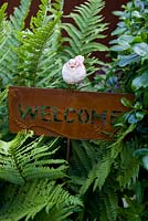 Little bird ornament perched on rusted steel welcome sign surrounded by fern foliage. 