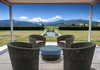 Rattan chairs and glass table with view to the mountains and vineyards beyond at Bhudevi Estate garden, Marlborough, New Zealand.
