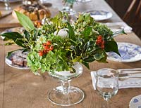 Footed glass candle centre piece arrangement made up of british grown flowers and foliage - hydrangea heads, viburnum davidii, holly and hips. Common Flower Farm, Somerset