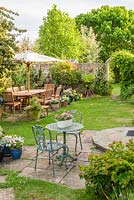 Outdoor dining area in secluded corner of family garden with wooden table, chairs and parasol. Small enamel trough wth dwarf antirrhinums on metal table.