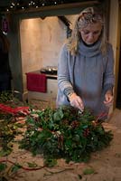 Christmas wreath making workshop.  Lady completing a wreath featuring Hedera - Ivy, red sprayed fir cones, dried apples, Pinus - Christmas tree twigs, red twigs and Cinnamon sticks. December, St Francis Cottage