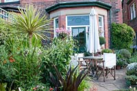 Patio with chairs and table at the back garden of a traditional Victorian red brick house. Planting of  Eucomis 'Sparkling Burgundy' Verbena  Yucca gloriosa 'Variegata', Lilium at Southlands,  July 