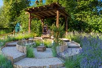The Lavender Garden, view of garden with its rustic rough-hewn timber gazebo, approached by a circular, stepped gravel pathway. In the centre are chairs and a crate serving as a table. Lavenders include Lavandula angustifolia and L. x intermedia cultivars. RHS Hampton Court Flower Show in 2016