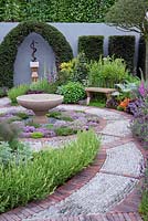 The St John's Hospice Garden, The Modern Apothecary.Brick and cobble stone path. Extensive planting of herbs around water basin in central circular bed. Oak seat in border and sculpture of Aesclepius on back wall. RHS chelsea Flower show 2016