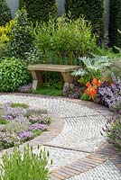 An oak wooden bench with living-willow-back, stone and brick circular paving and planting of Lavendula 'Helmsdale' and Thymus. The St John's Hospice - A Modern Apothecary. RHS Chelsea Flower Show 2016. Designer: Jekka McVicar, Sponsor: St John's Hospice. Silver-Gilt