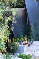 The Telegraph Garden, view of a fire bowl, irregular geometric bronze fin sculptures, seating area with stone benches surrounded by Isoplexus canariensis - Canary Island Foxgloves, Maytenus boaria, Schinus molle. RHS Chelsea Flower Show 2016. Designer: Andy Sturgeon - Sponsor: The Telegraph
