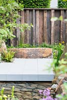 The M and G Garden, view of sawn stone path over a pond with clear water and stone raised bed in front of wooden fence surrounded by Blechnum spicant, Iris Pseudoacorus and Quercus pubescens. RHS Chelsea Flower Show, 2016 Designer: Cleve West MSGD, Sponsor: M and G 