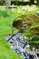 A pair of zig-zag perching sticks are wedged into pebbles beside the still pool.