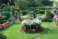 Colourful mixed beds filled shrubs, perennials and tender bedding plants, including Lobelia, Calendula, Begonia, Clematis, Achillea, Leucanthemum and Rosa with path across the lawn to pergola.