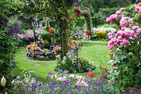 View of colourful small back garden with borders filled with tender bedding plants, perennials, Rosa 'Pomponella' and Clematis on trellis and pond 