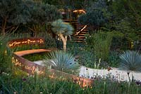 The Winton Beauty of Mathematics Garden lit at night, RHS Chelsea Flower Show 2016. Illuminated mathematical symbols cut into band of copper running through the garden forming back of bench and bannister for staircase. Pinus sylvestris 'Glauca' - blue Scot's pine, Yucca rostrata - Beaked Yucca