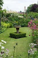 A front garden lawn with sundial and topiary box, surrounded by a mixed perennial border with cottage garden plants.
