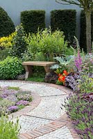 A Modern Apothecary. A brick and cobble path leading to an oak bench is edged in French lavender, Lavandula stoechas 'Helmsdale', thyme, nasturtium and foxglove. Designer: Jekka McVicar. RHS Chelsea Flower Show 2016