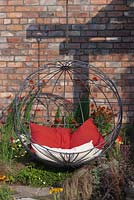 A contemporary spherical suspended metal chair against a red brick wall with perennials in hot colours -   July, Tatton Park RHS Flower Show 2014